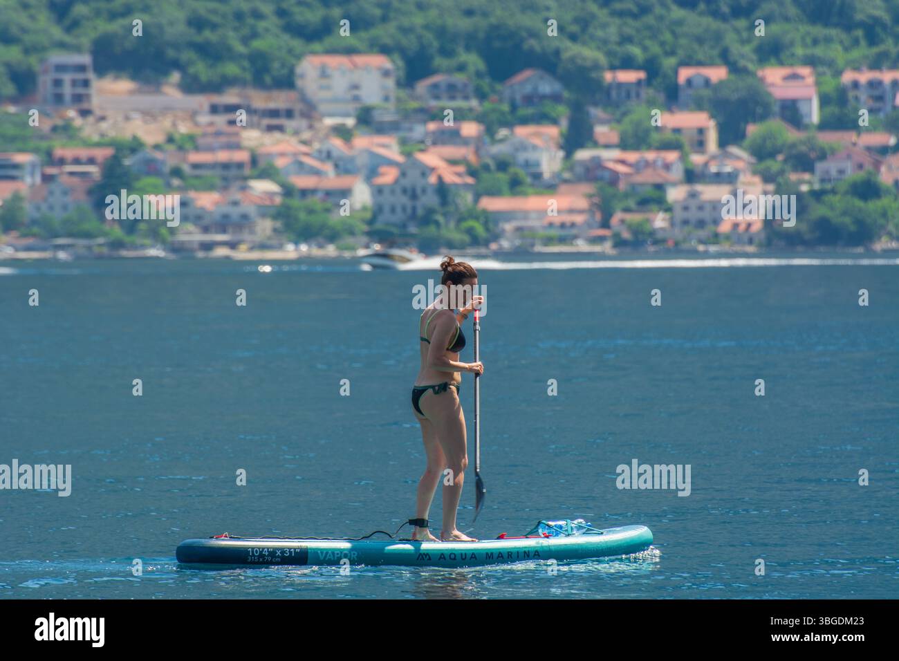 Junge Frau Standup Paddleboarding (SUP) in Kotor Bay, Montenegro mit traditionellem montenegrinischen Dorf im Hintergrund. Stockfoto