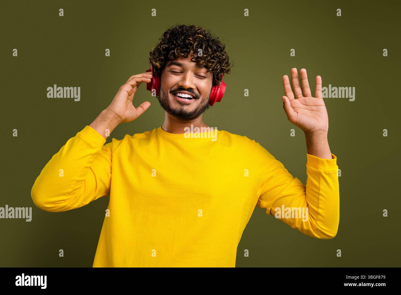 Fröhlicher junger Mann, der Musik über Kopfhörer hört, während er lächelt und gespendet, in einem leuchtend gelben Hemd und lässigem Stil Stockfoto