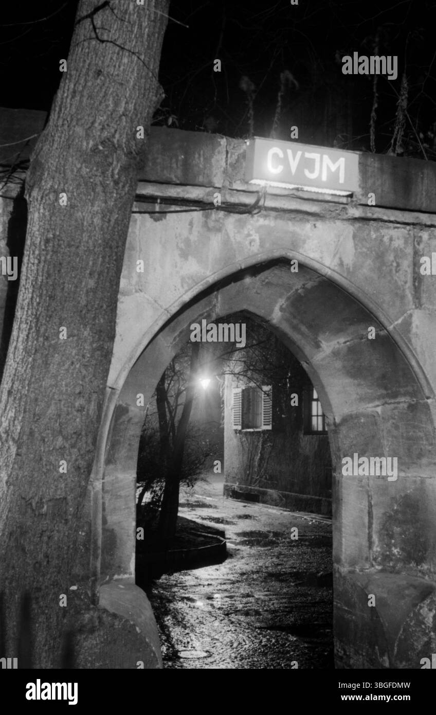 Nächtliche Ansicht eines historischen Torbogens mit beleuchtetem CVJM-Schild, CVJM ist ein christlicher Verein junger Männer. Blick durch den Eingangstorbogen zum CVJM-Verein auf eine regennasse, gepflasterte Gasse mit Straßenlaterne und Gebäude im Hintergrund, um 1955. Stockfoto