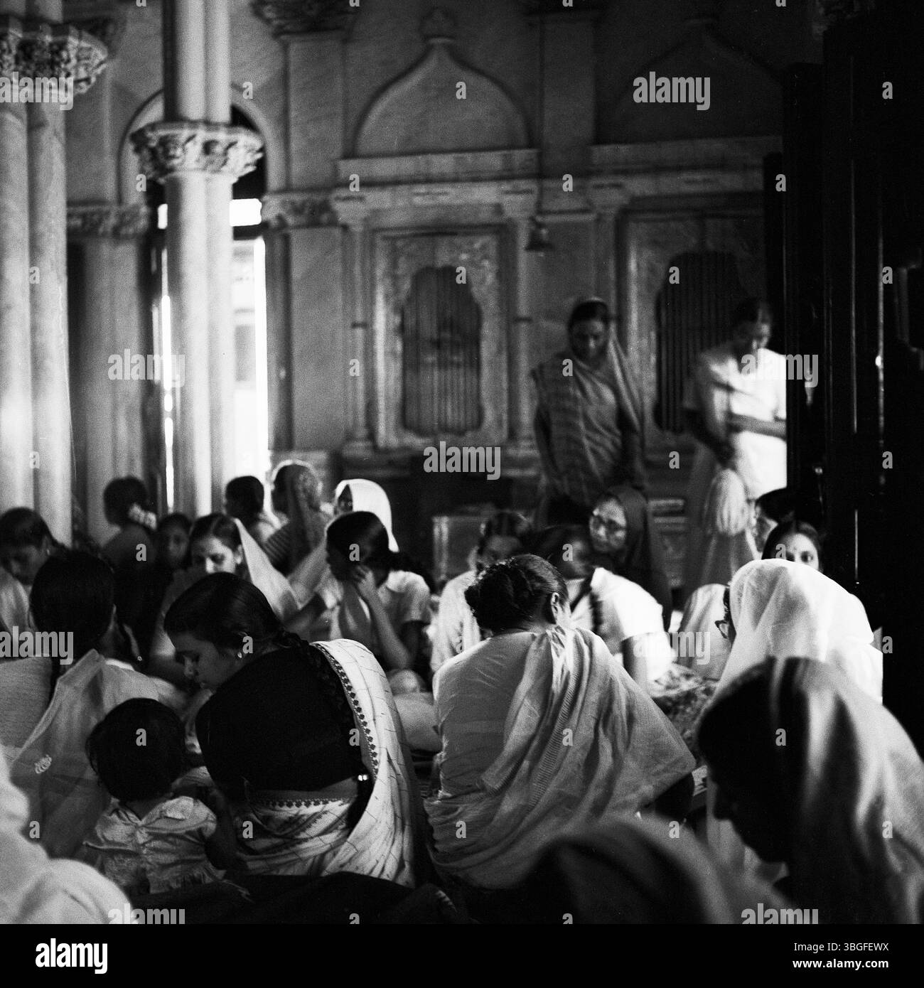 Indien, um 1963: Frauen in traditionellen Saris sitzen dicht gedrängt auf dem Boden eines reiches verzierten Raumes, vermutlich zum Gebet, zur Meditation oder für eine gemeinschaftliche Veranstaltung. Zwei Frauen stehen im Hintergrund, das Licht fällt durch Fensterbögen und beleuchtet dezent das geschehen. Stockfoto