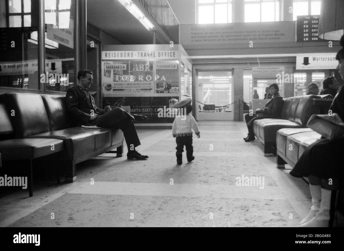 Ein kleines Kind mit übergroßer Uniformmütze steht auf dem Boden der Abflughalle im Flughafen München-Riem und blickt zu zwei US-amerikanischen Soldaten, die entspannt auf den Ledersesseln der Wartezone sitzen. Stockfoto