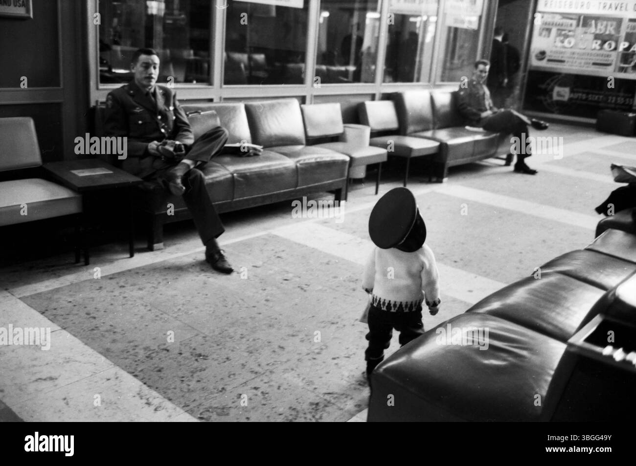 Ein kleines Kind mit übergroßer Uniformmütze steht auf dem Boden der Abflughalle im Flughafen München-Riem und blickt zu zwei US-amerikanischen Soldaten, die entspannt auf den Ledersesseln der Wartezone sitzen. Die Szene wirkt wie ein stiller Moment des neugierigen Beobachtens – zwischen militärischer Präsenz und kindlicher Unbefangenheit. Stockfoto