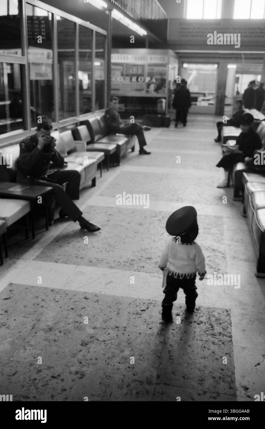 Ein kleines Kind mit übergroßer Uniformmütze steht auf dem Boden der Abflughalle im Flughafen München-Riem und blickt zu zwei US-amerikanischen Soldaten, die entspannt auf den Ledersesseln der Wartezone sitzen. Stockfoto
