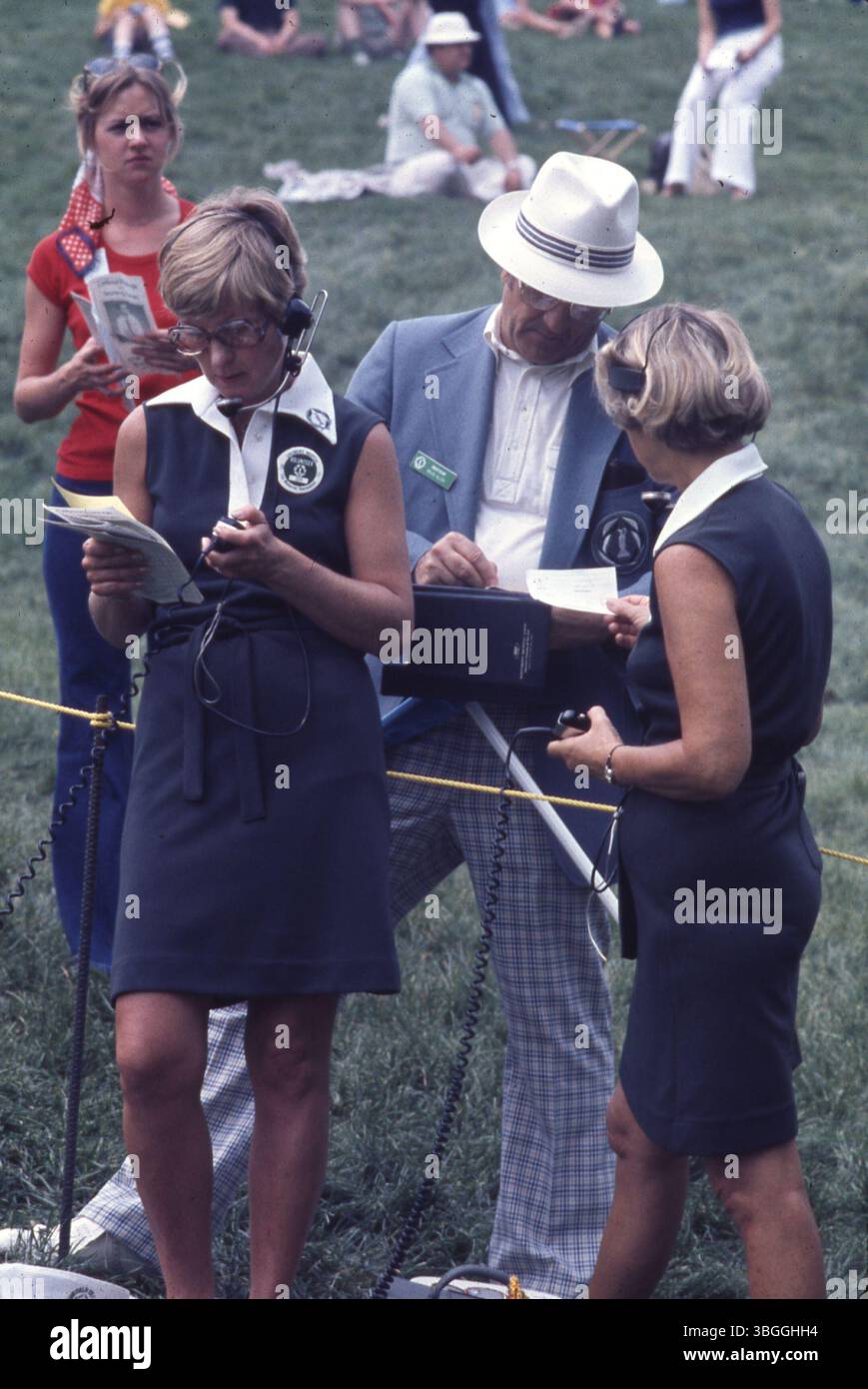Beim Memorial Tournament 1977 helfen drei Freiwillige des Muirfield Village Golf Club. Zwei Frauen tragen passende Kleider und Kopfhörer, während ein Mann mit weißkrempigem Hut etwas schreibt. Eine weibliche Zuschauerin in der Nähe hält ein Programm ab. Stockfoto