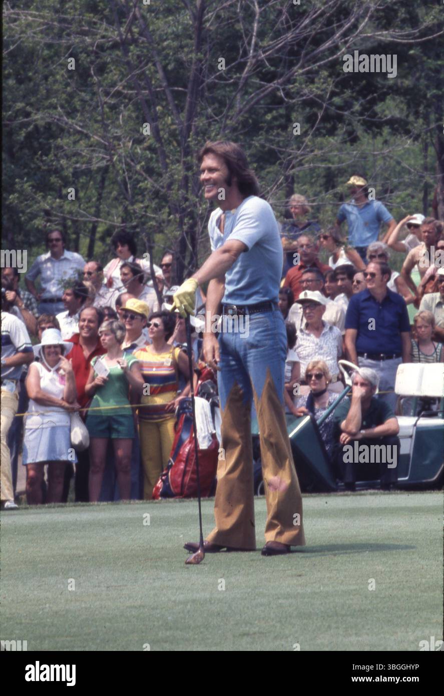 Der Musiker Glen Campbell steht bei einem Abschlag mit einem Holzclub während des Memorial Tournament pro-am 1977 im Muirfield Village Golf Club. Die Zuschauer beobachten, wie er mit Gerald Ford, Jack Nicklaus, Roger Maltbie, Jackie Gleason und Bob Hope spielt. Stockfoto
