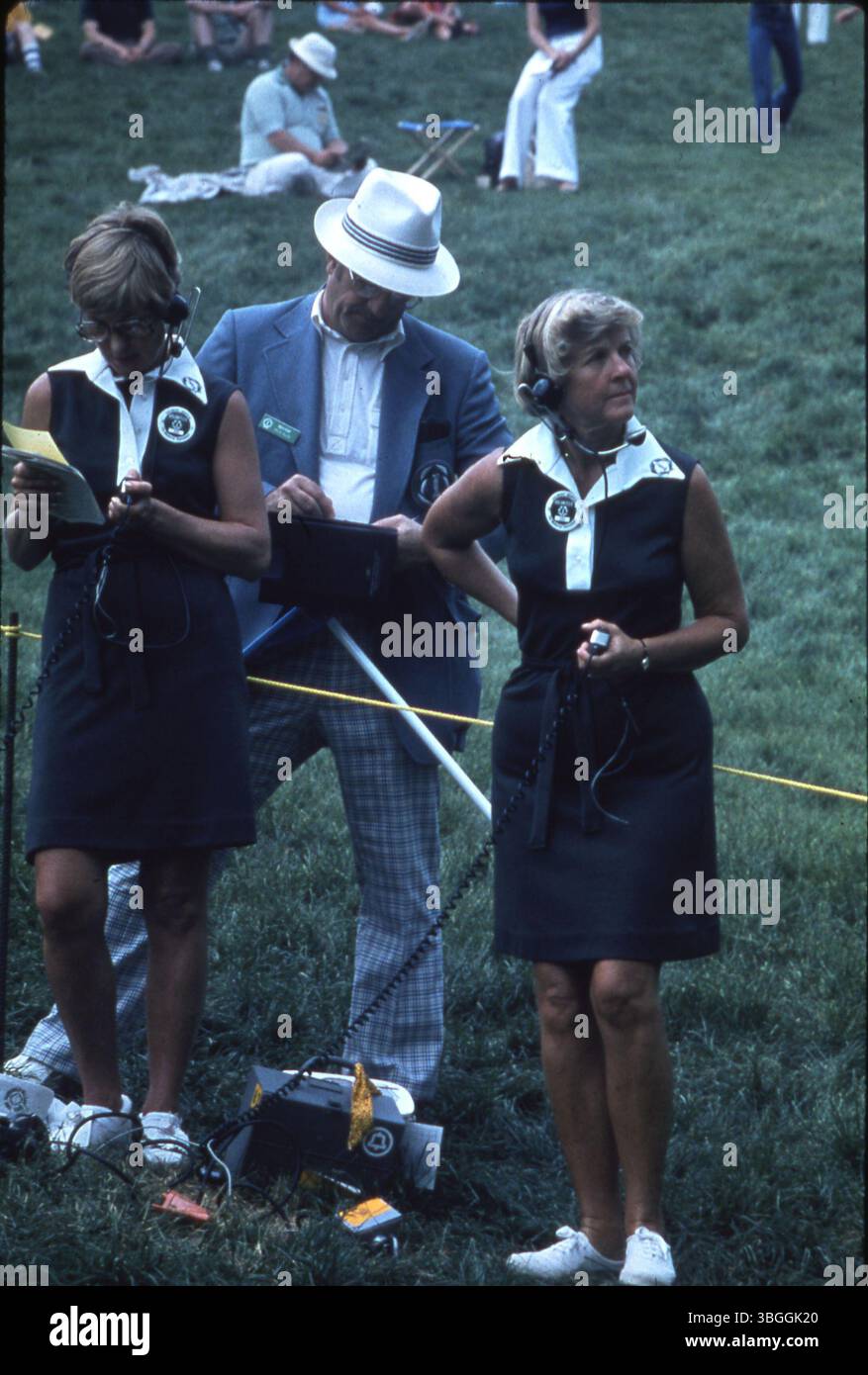Drei Freiwillige beim Memorial Tournament 1977 im Muirfield Village Golf Club. Zwei Frauen in passenden Kleidern mit Kopfhörern und ein Freiwilliger mit weißem Schirm unterstützen die Veranstaltung. Die Zuschauer sind in der Ferne zu sehen. Stockfoto