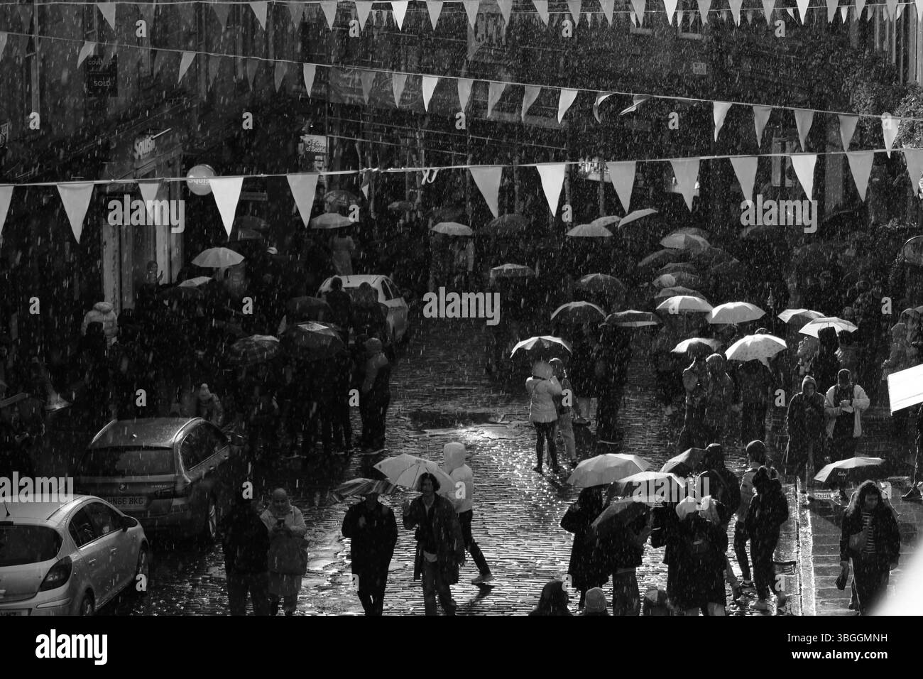 Menschenmassen mit Sonnenschirmen wandern im Regen auf der Victoria Street, die von Licht auf nassen Kopfsteinpflastersteinen in einer dramatischen schwarz-weißen Straßenszene umrahmt wird. Stockfoto