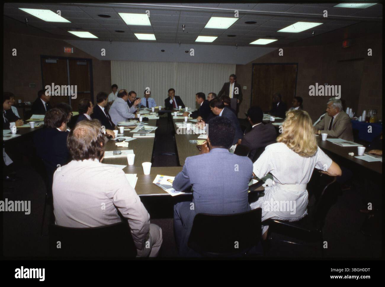 Eine Gruppe von Männern und Frauen sitzt an langen Tischen während eines nicht identifizierten Treffens. Eine Person steht und spricht, während der Rest aufmerksam zuhört. Stockfoto