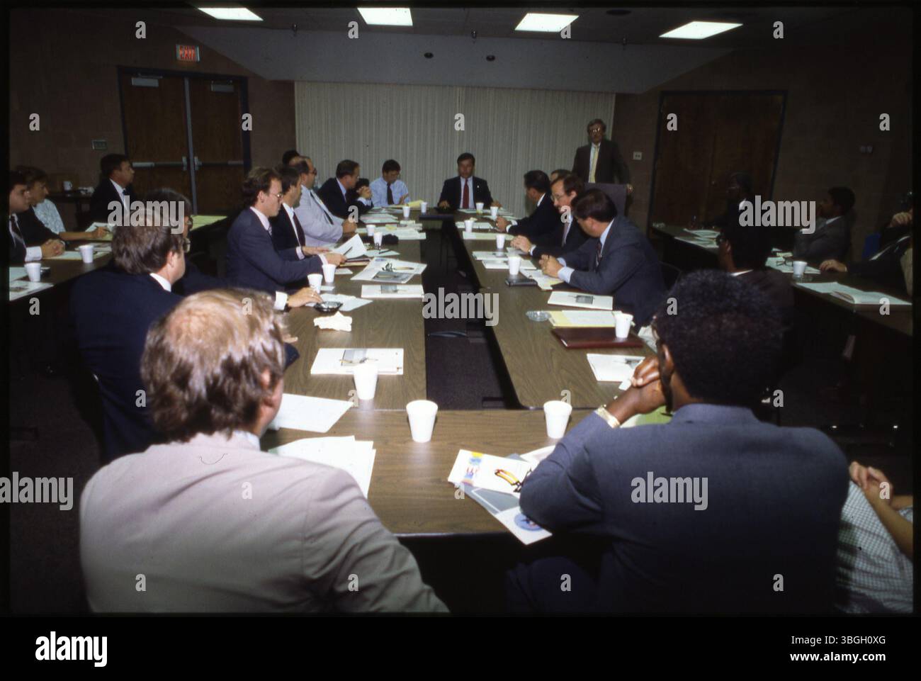 Eine Gruppe von Männern, die an langen Tischen sitzen, mit einer Person, die steht und spricht. Der Tatort scheint von einem nicht identifizierten Treffen zu stammen, das zwischen 1974 und 1979 stattfand. Die Einzelpersonen hören aufmerksam zu, wenn eine Person die Gruppe anspricht. Stockfoto
