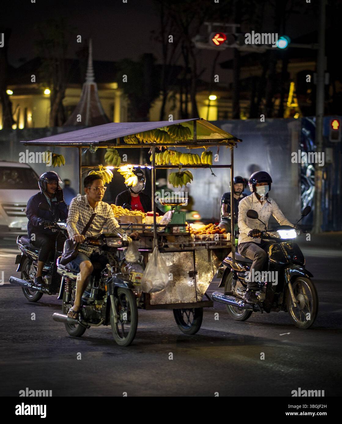 Eine geschäftige Nachtszene mit einem Straßenverkäufer, der Bananen aus einem Wagen verkauft, umgeben von Motorrädern und Stadtlichtern. Die Atmosphäre ist lebhaft und man Stockfoto