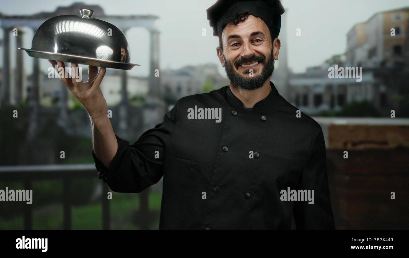 Der Koch mit Bart in schwarzer Uniform hält eine silberne Platte, die in der Altstadt roms lächelnd mit antiker Architektur im Hintergrund, die kulinarische kunst zeigt Stockfoto