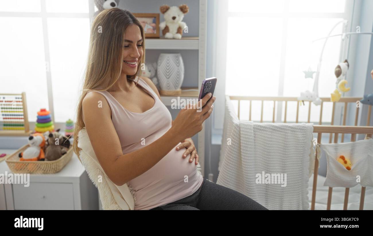 Schwangere Frau mit langen blonden Haaren sitzt auf dem Bett in einem gemütlichen Schlafzimmer und lächelt an ihrem Telefon, umgeben von Babyspielzeug und einer Wiege, was eine warme Familie schafft Stockfoto