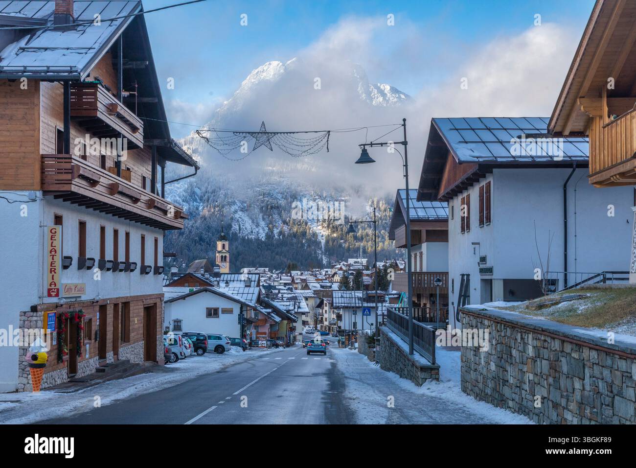 Winterszene in Sappada säumen alpine Häuser und Geschäfte die Hauptstraße mit einem Dolomitengipfel im Hintergrund, Provinz Udine, Dolomiten, Italien Stockfoto
