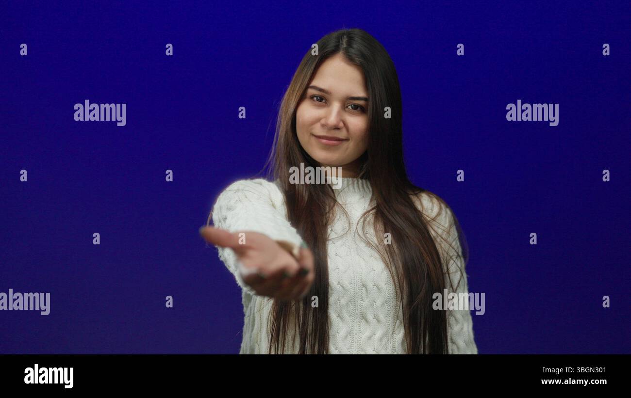 Frau, die über blauem isoliertem Hintergrund zur Kamera greift, trägt einen Pullover mit langen Haaren und einladendem Ausdruck Stockfoto