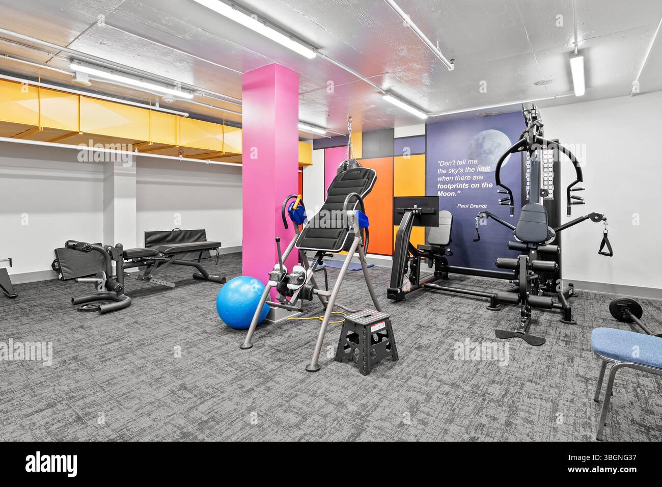 Fitnessstudio mit rosa Wänden und blauem Ball. Der Fitnessraum ist voll mit Fitnessgeräten und einem Zitat an der Wand Stockfoto