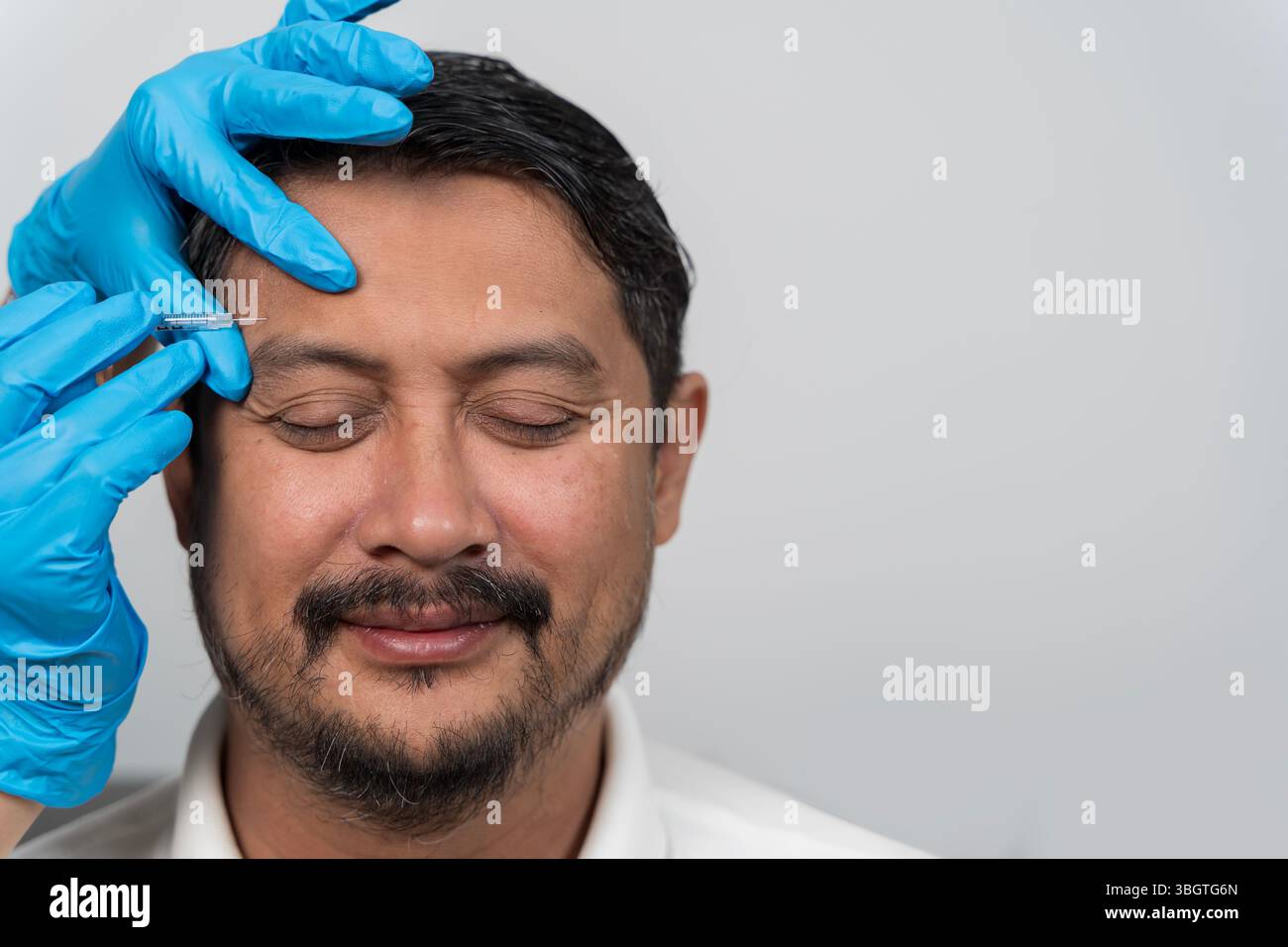 Lächelnder Mann mittleren Alters, der kosmetische Injektion in der Nähe der Augenpartie erhält. Ästhetische Behandlung durch einen Profi mit blauen Handschuhen. Konzept der Hautpflege Stockfoto