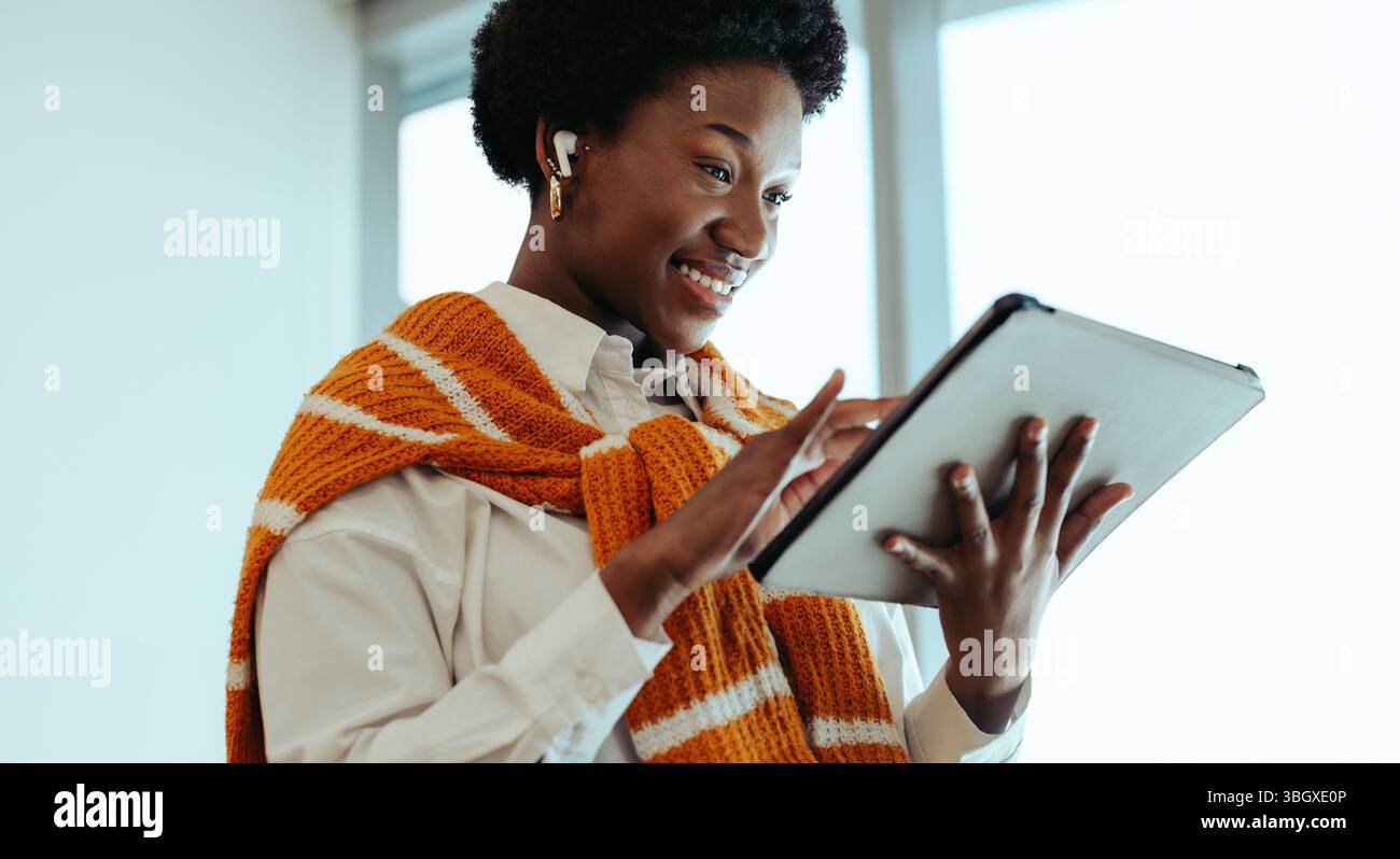 Eine Frau lächelt, während sie ein digitales Tablet in einem modernen Büroumfeld verwendet. Sie trägt einen leuchtenden, orange gestreiften Pullover und schnurlose Ohrstöpsel, um zu schaffen Stockfoto