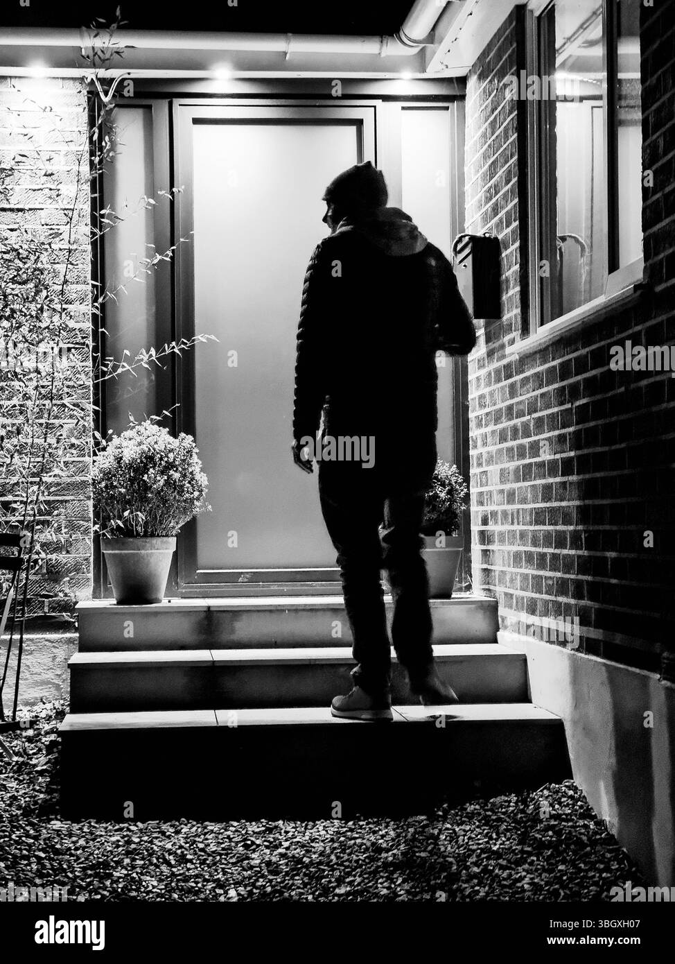 Schwarz-weiße Silhouette einer Person, die nachts vor einem Haus steht, beleuchtet von der Veranda Licht - Krimi-Einbruchthriller-Buchcover-Konzept Stockfoto