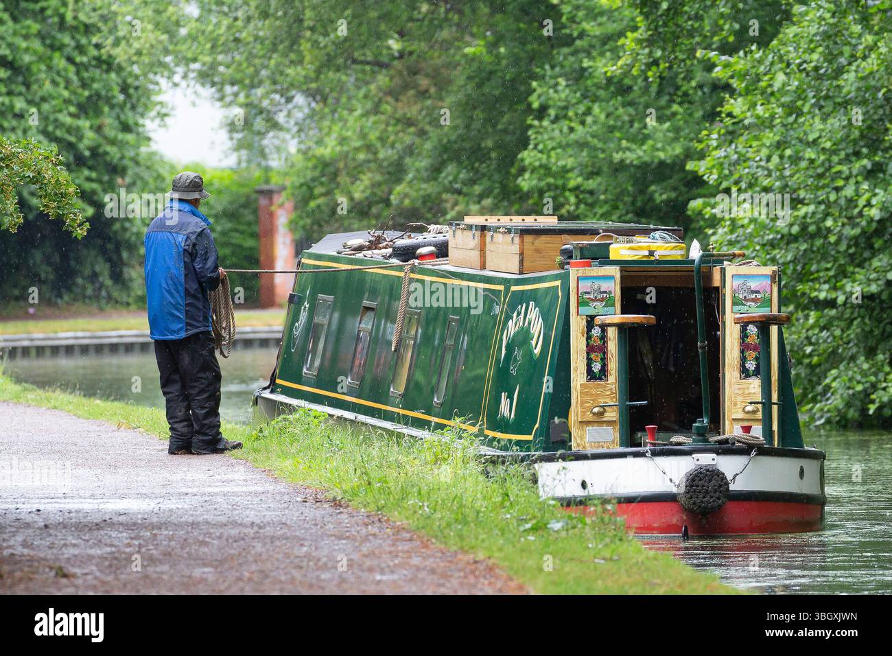 Rückansicht eines britischen Mannes in wasserdichtem Mantel, der im Sommerregen sein Schmalboot mit einem Seil an die Anlegestellen am Wasser an einem Kanal von Worcestershire zieht. Stockfoto