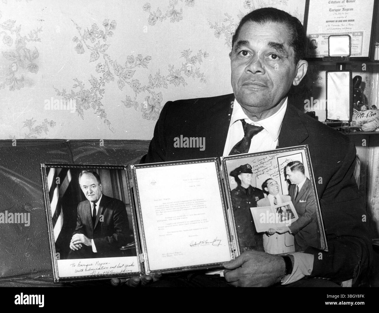 Enrique Negron, ein Ex-Lebensmittelhändler aus Bronx, der einem Polizisten half, der von einem wütenden Mob belagert wurde, hält ein Bild und eine Laudatio von Vizepräsident Hubert H. Humphrey und ein Foto von sich selbst mit PTL. Philip Siegel, der Mann, dem er half, und sein Abgeordneter Jim Scheuer, während er über seine Zukunft in seinem Haus nachdenkt. - 17. Januar 1966 Stockfoto