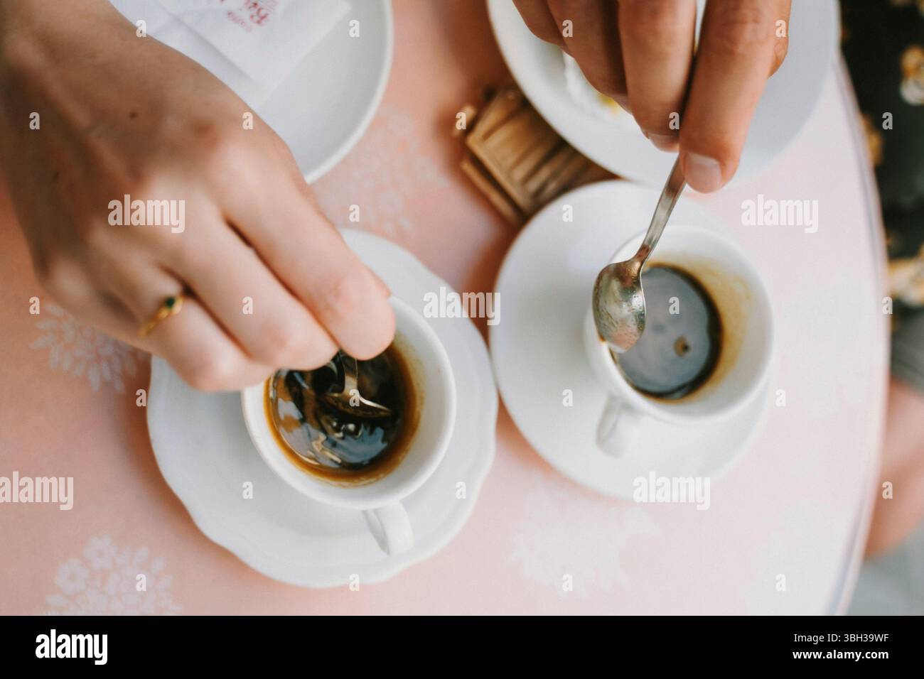 Zwei Personen genießen einen ruhigen Moment im Café und rühren Kaffee in weißen Tassen um. Die Nahaufnahme fängt die Hände mitten in der Action ein und betont Entspannung und Mitgefühl Stockfoto