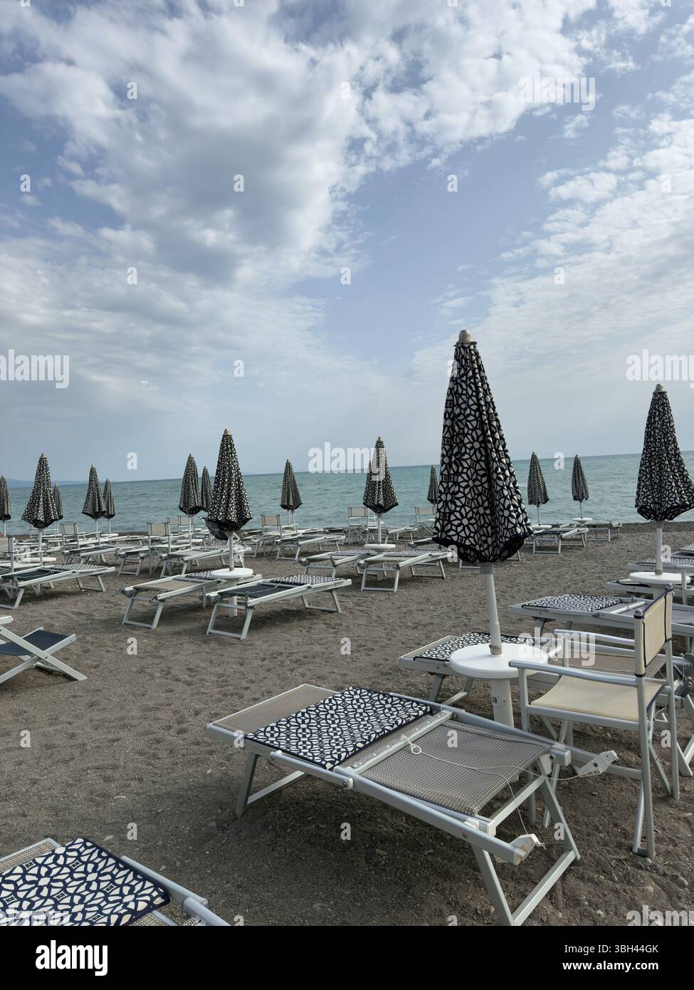 Geschlossene Sonnenschirme und Sonnenliegen am Morgenstrand vor dem Hintergrund der Küste - Smartphone-aufgenommenes Stockfoto