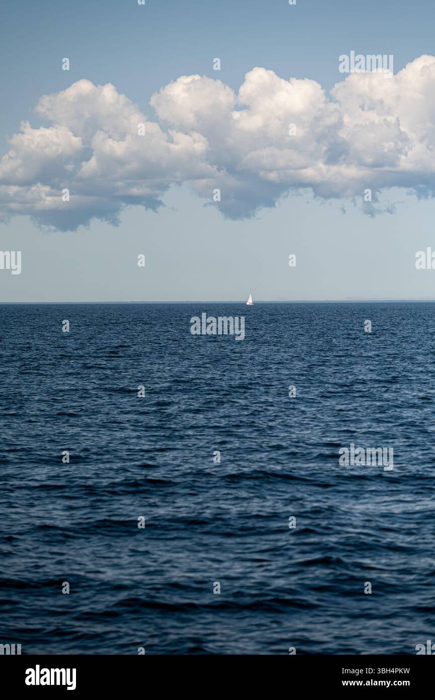 Ein kleines Segelboot segelt friedlich über das weitläufige blaue Wasser unter einem wolkenreichen Himmel. Stockfoto