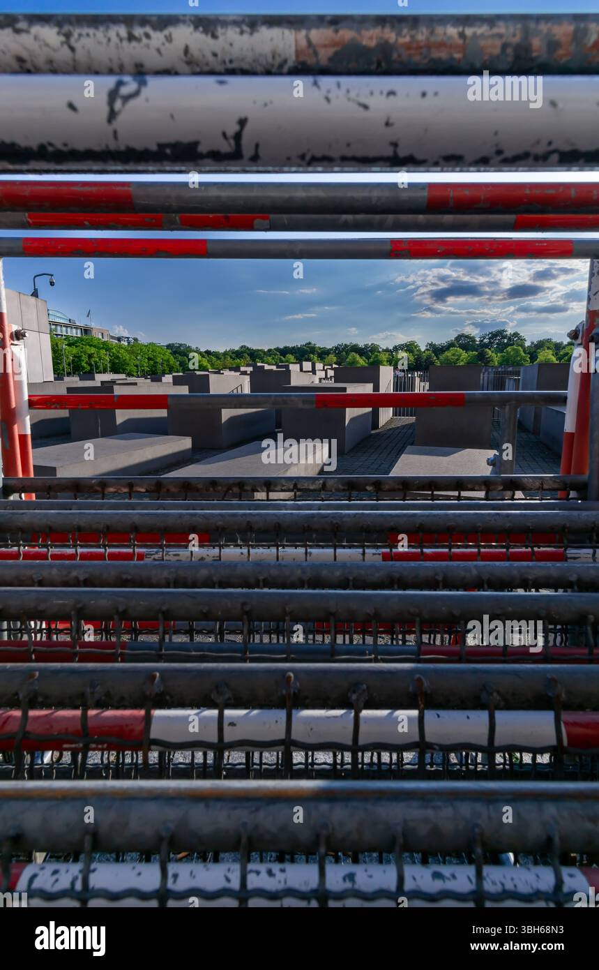 Das Berliner Holocaust-Mahnmal wird durch gestapelte rot-weiße Barrieren gesehen und bildet einen mehrschichtigen visuellen Rahmen im vertikalen Format. Stockfoto