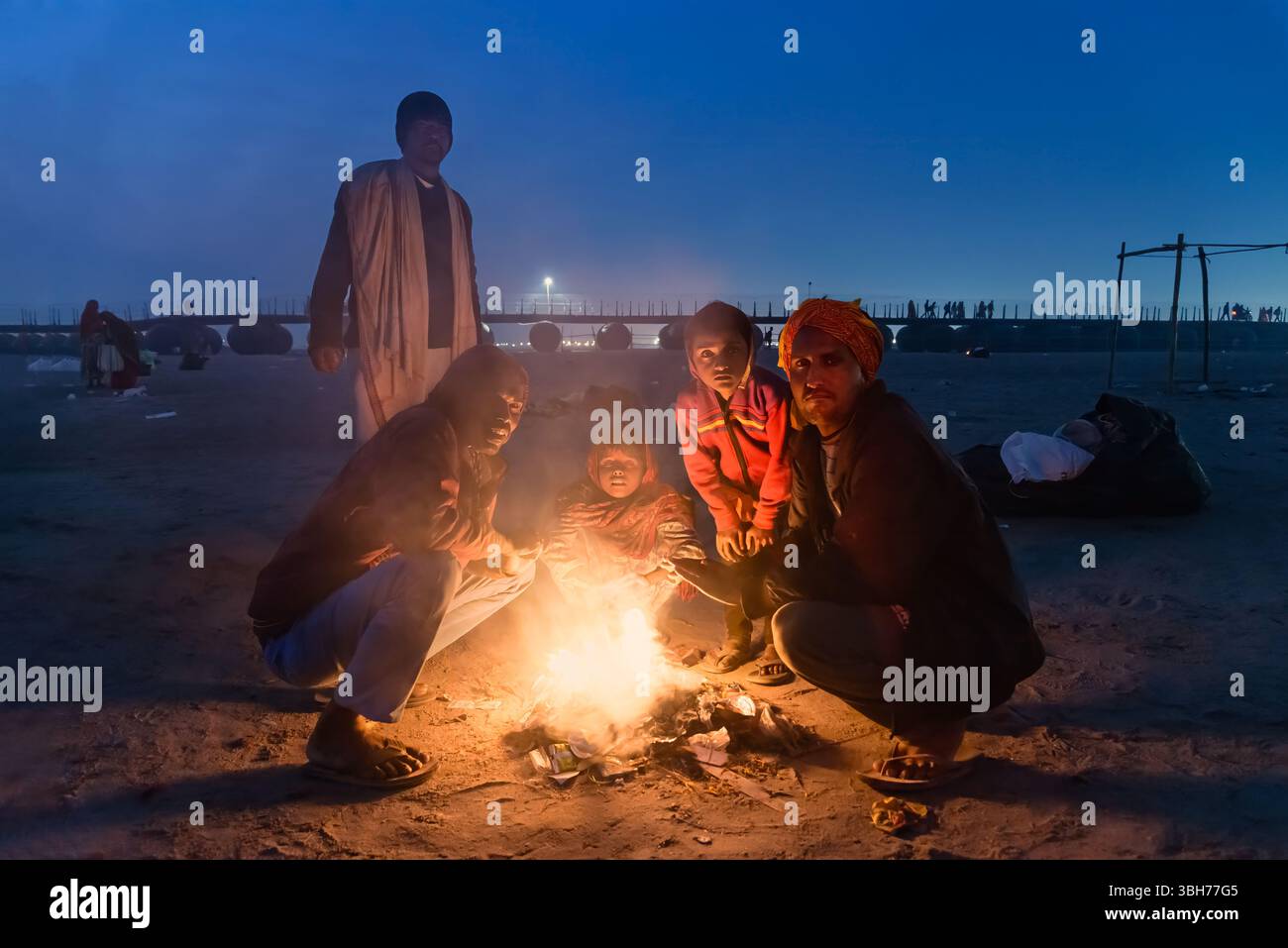 Prayagraj, Indien - 26. Februar 2025: Eine Gruppe von Gläubigen sitzt bei Sonnenuntergang an einem Sandufer während der Kumbh Mela in Indien um ein kleines Feuer. Stockfoto