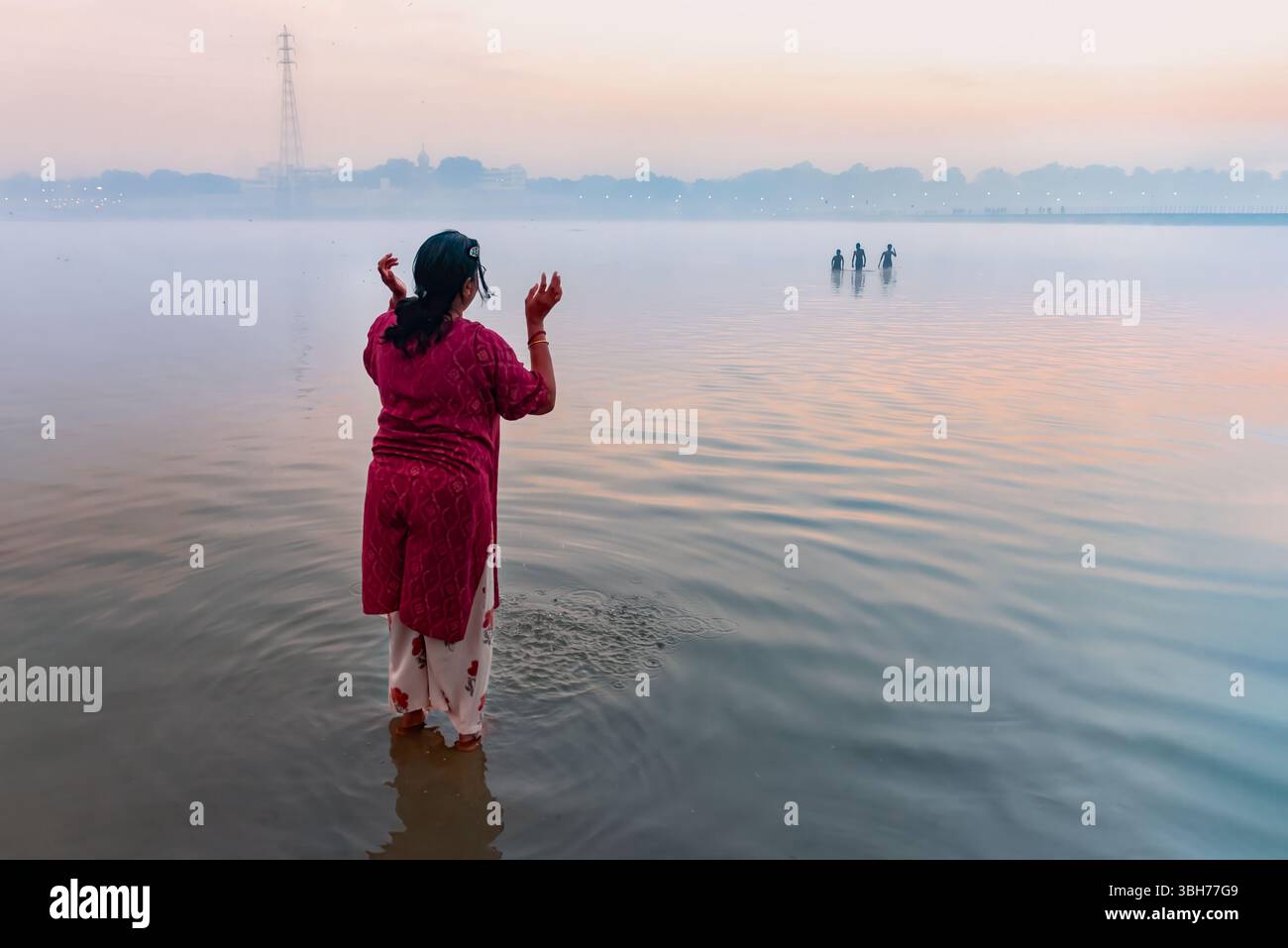 Prayagraj, Indien - 26. Februar 2025: Frau steht hüfttief im Fluss bei Sonnenaufgang während Kumbh Mela, Indien, mit erhobenen Händen im Gebet. Stockfoto
