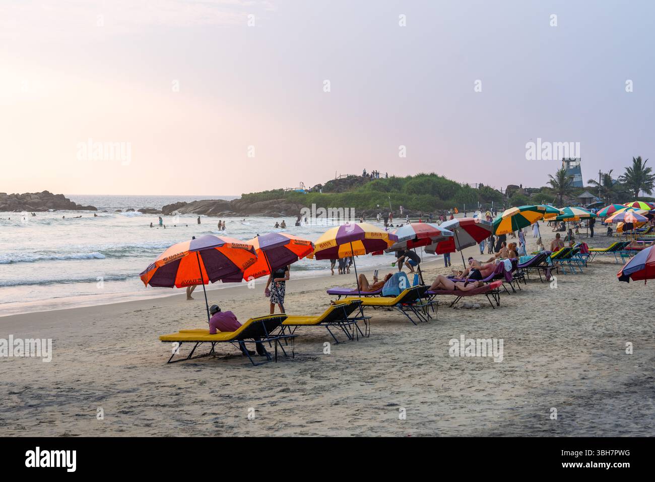 Touristen und Einheimische entspannen sich unter bunten Sonnenschirmen am Kovalam Beach bei Sonnenuntergang, Kerala, Südindien Stockfoto