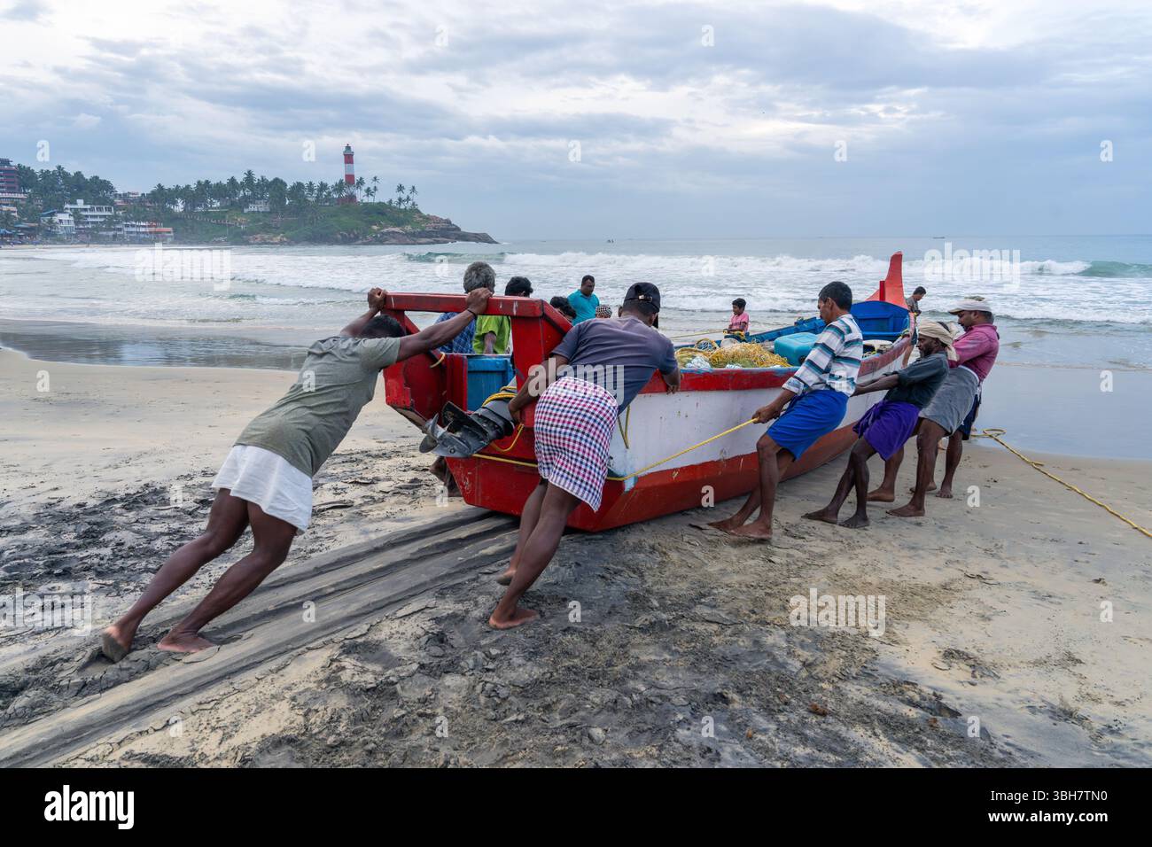 Traditionelle Fischer ziehen ein rotes Fischerboot an den Strand in Kovalam, Kerala, Indien, mit dem berühmten Leuchtturm in der Ferne Stockfoto