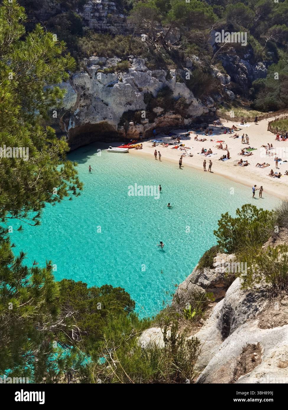 Türkisfarbener Strand, weißer Sand, mediterrane Klippen - Cala Macarelleta in Menorca, Spanien, von oben gesehen mit lebhaftem Wasser, Kiefernwald und Menschen - Smartphone-aufgenommenes Stockfoto