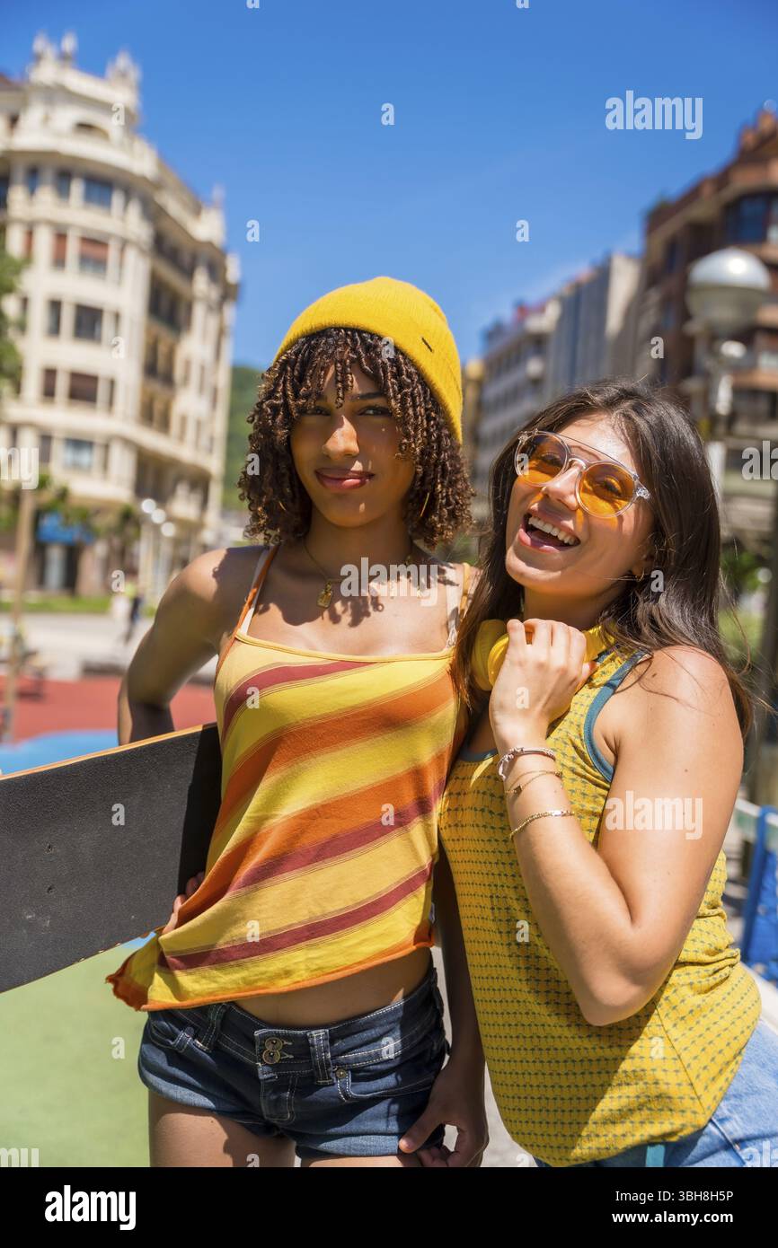 Zwei Freundinnen, die einen sonnigen Tag in der Stadt genießen und ihre Jugendliche Energie und ihren urbanen Stil mit Skateboard und Kopfhörern unter Beweis stellen Stockfoto