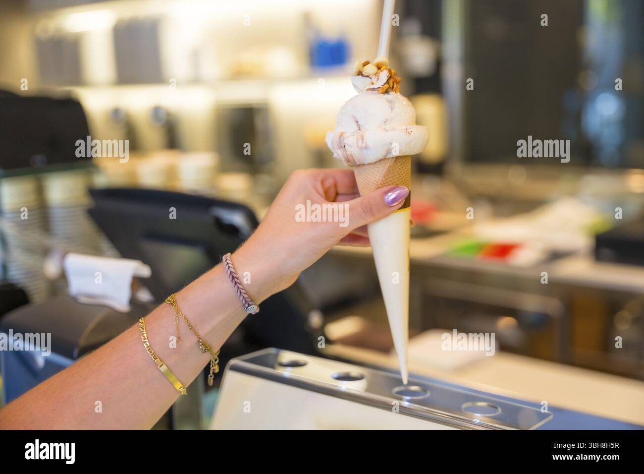 Frauen Hand mit Armbändern, die einen köstlichen Eiskegel mit knusprigen Nüssen halten, genießen eine Leckerei in einer Gelateria Stockfoto