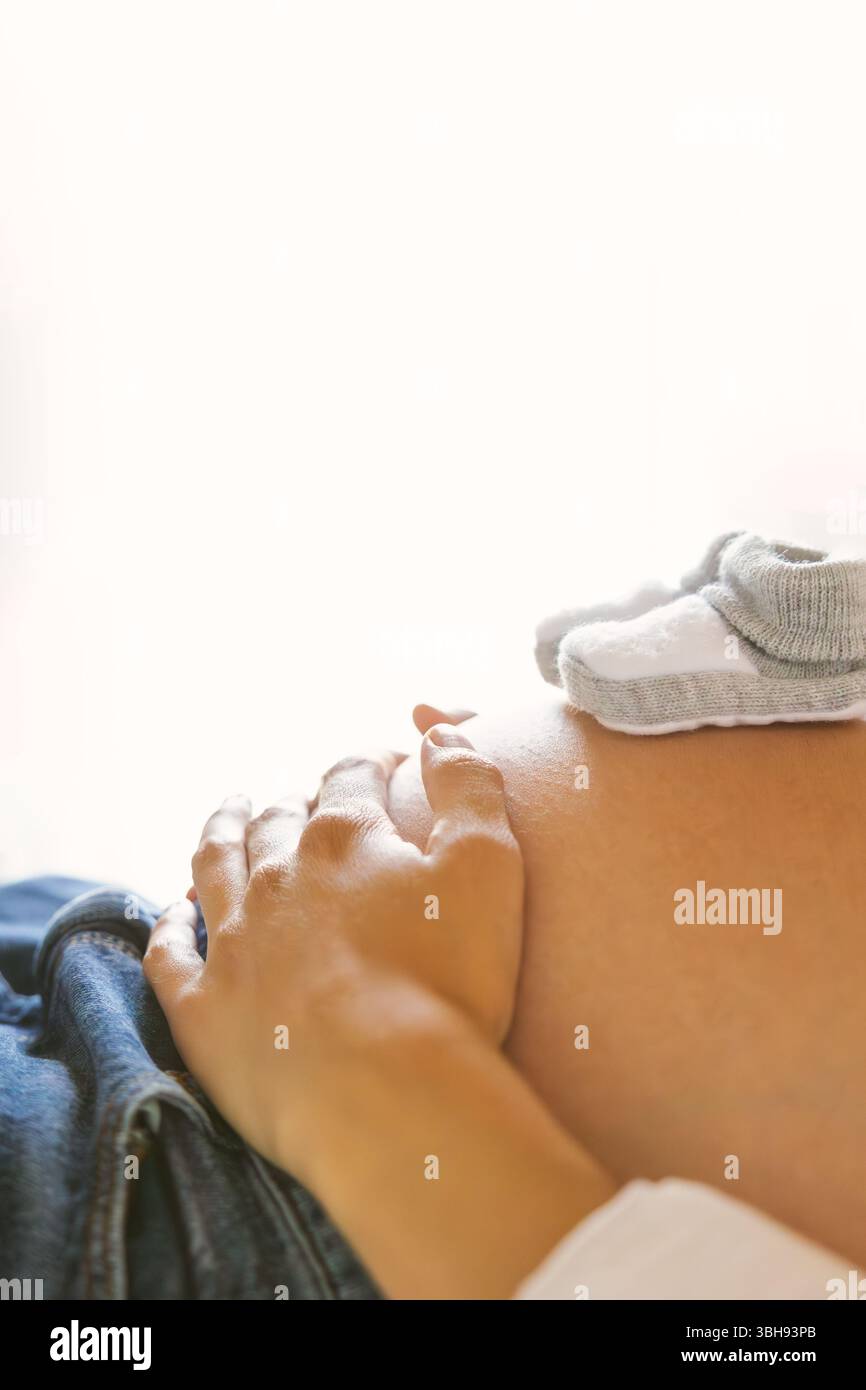 Hand am schwangeren Bauch mit Babyschuhen in sanfter Naturbeleuchtung. Lifestyle. Stockfoto