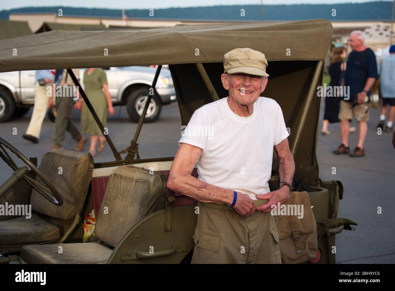 Ein älterer Mann in einem weißen T-Shirt und khakifarbenen Hosen steht neben einem alten Militärjeep. Er trägt eine khakifarbene Mütze und ein blaues Armband. Der Jeep ist oliv Stockfoto