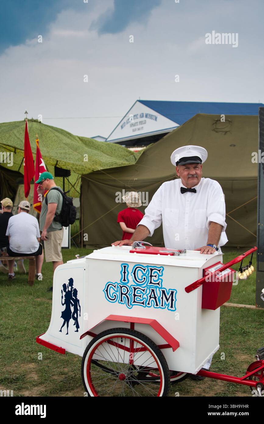 Ein Eisverkäufer in weißer Uniform steht neben seinem Eiswagen bei einer Veranstaltung im Freien. Der Wagen ist weiß mit roten Akzenten und der Aufschrift „EIS“ Stockfoto