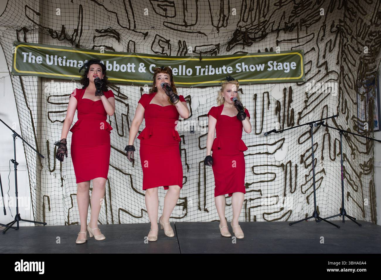 Drei Frauen in roten Kleidern singen auf einer Bühne mit Camouflage-Muster. Ein Banner oben lautet: „The Ultimate Abbott & Costello Tribute Show St Stockfoto