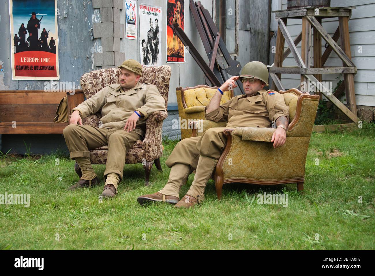 Zwei Männer in Uniformen der US-Armee sitzen draußen in Sesseln. Im Hintergrund stehen antike Holzmöbel und Propagandaplakate. Die Einstellung Stockfoto