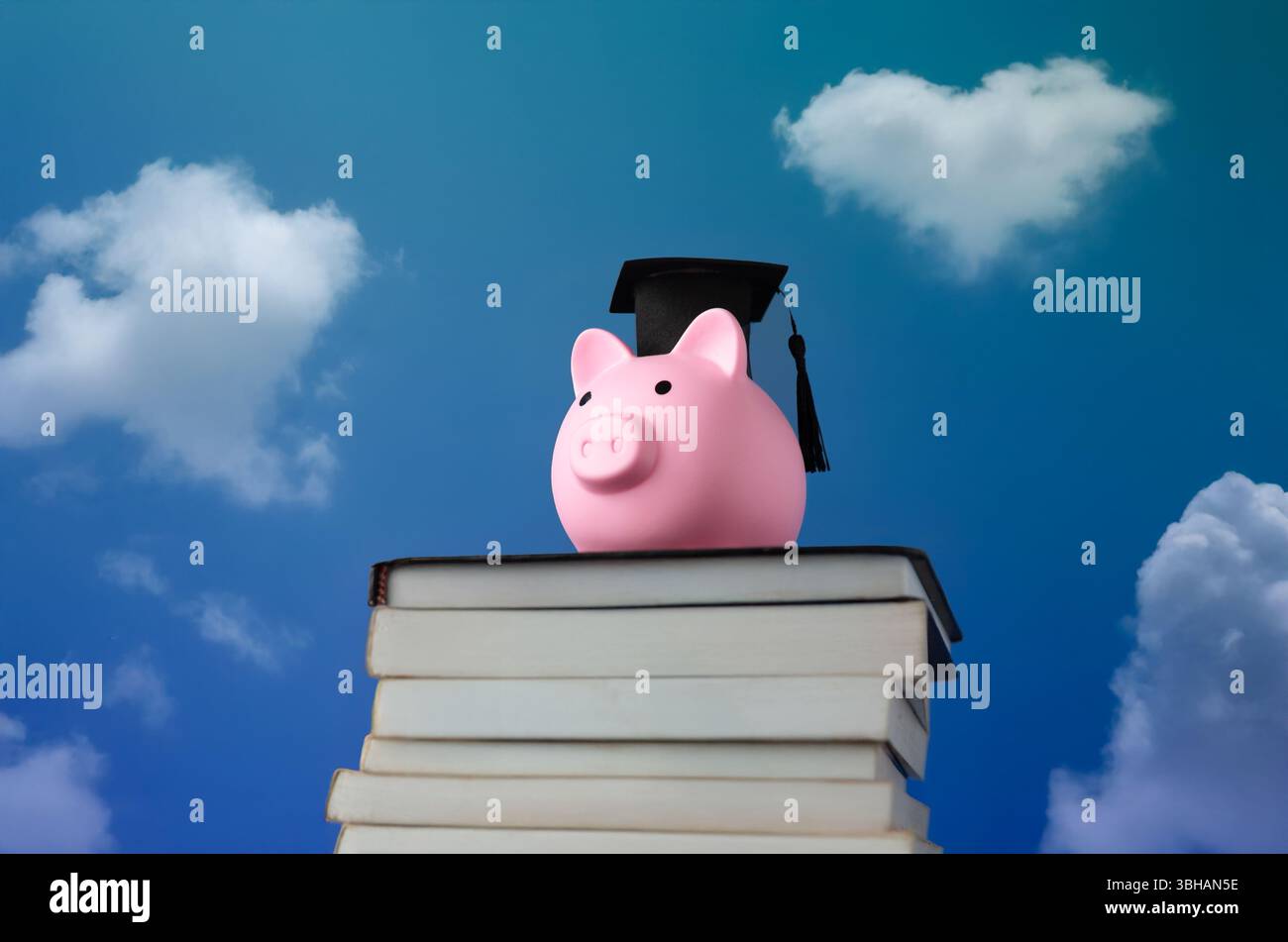 Eine pinkfarbene Schweinebank mit einer schwarzen Graduierungsmütze auf einem Stapel Bücher vor einem hellblauen Himmel mit Wolken. Akademische Leistungen Stockfoto