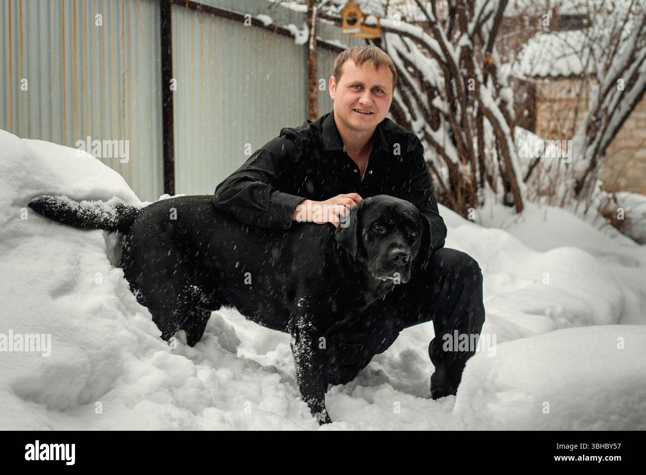 Ein Mann und ein schwarzer Labrador Retriever spielen im Neuschnee Stockfoto