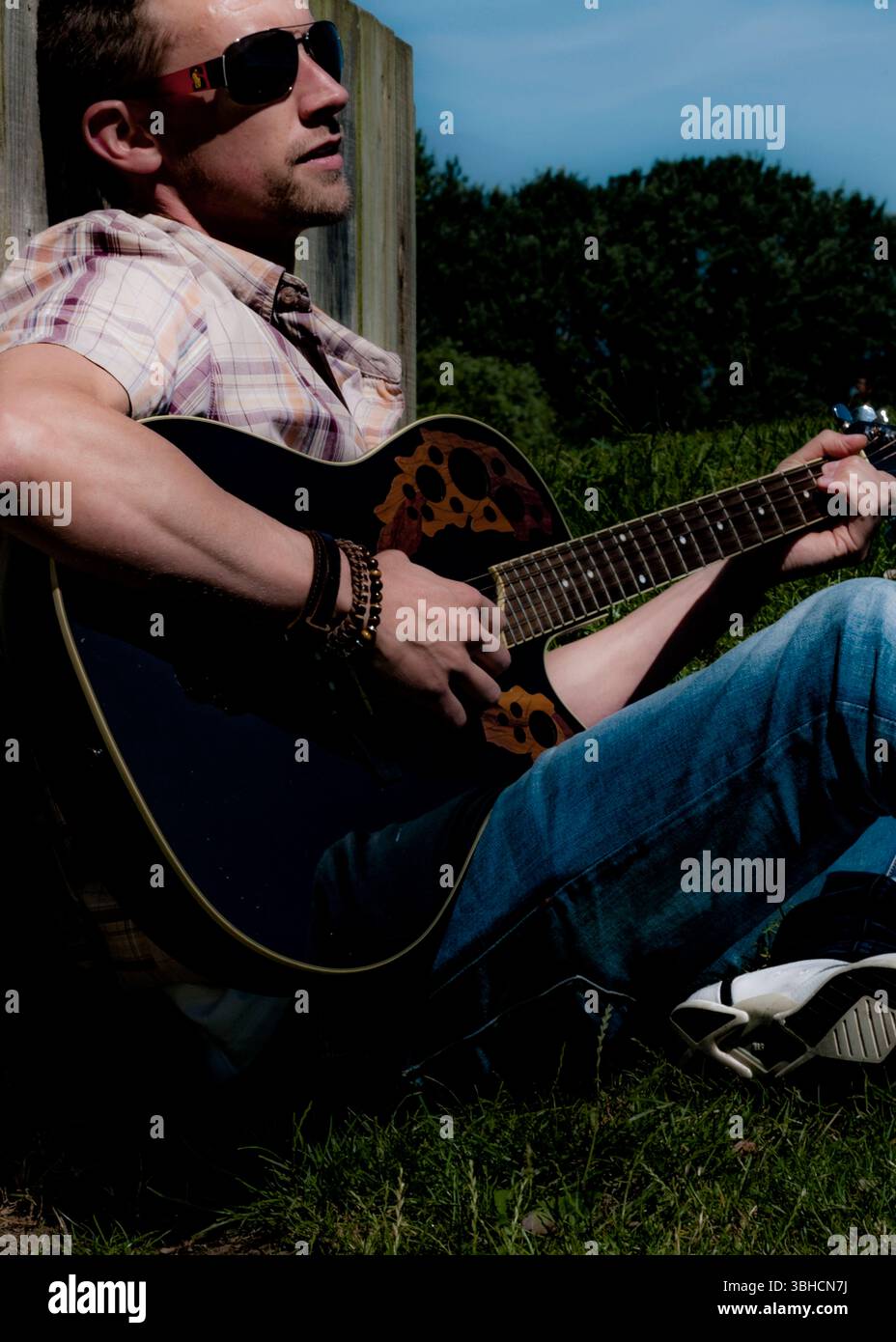 Ein Mann mit Sonnenbrille, kariertem Kurzarm-Hemd, blauer Jeans und weißen Sneakers sitzt vor einem Holzzaun und spielt eine schwarze Akustikgitarre. Er Stockfoto