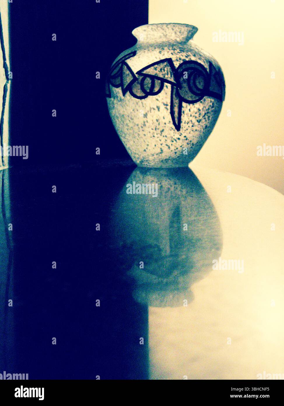 Auf einer reflektierenden Oberfläche befindet sich eine weiße Vase mit dunklen geometrischen Mustern. Der Hintergrund wird zwischen einem dunklen und einem hellen Bereich aufgeteilt, wodurch entsteht Stockfoto