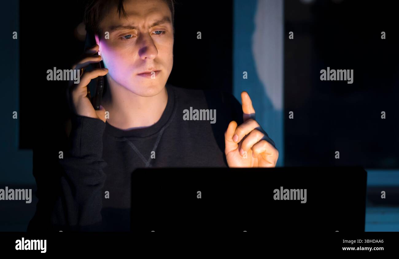 Ein junger Mann arbeitet online auf einem Laptop und spricht nachts mit Kunden oder technischem Support über ein Mobiltelefon, ein erfolgreicher Selbstständiger, intensiv Stockfoto