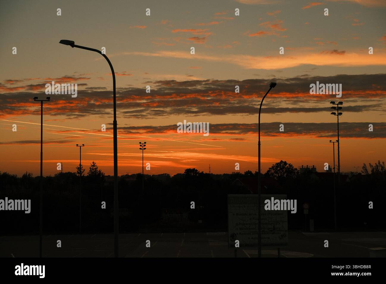 Urbane Sonnenuntergangsszene mit Silhouetten aus Straßenlaternen und Bäumen vor einem dramatischen Himmel, der in warmen Orange- und Rottönen gemalt ist. Stockfoto
