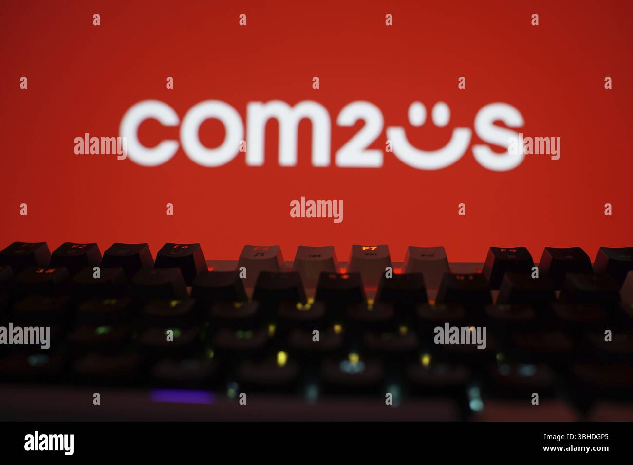 Com2us-Logo auf rotem Hintergrund mit einer Silhouettentastatur mit farbenfrohen Lichtern Stockfoto