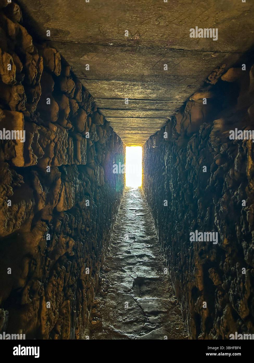 Schmaler Steintunnel mit rauen Wänden und Decke, der zu einem hellen Licht am Ende führt Ein dramatischer und geheimnisvoller Durchgang. - Smartphone-aufgenommenes Stockfoto