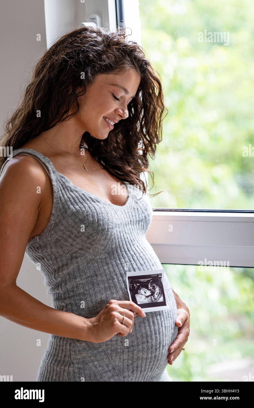 Hübsche Frau, die ihren schwangeren Bauch mit Freude ansieht, während sie das Ultraschallbild des Babys neben sich hält Stockfoto