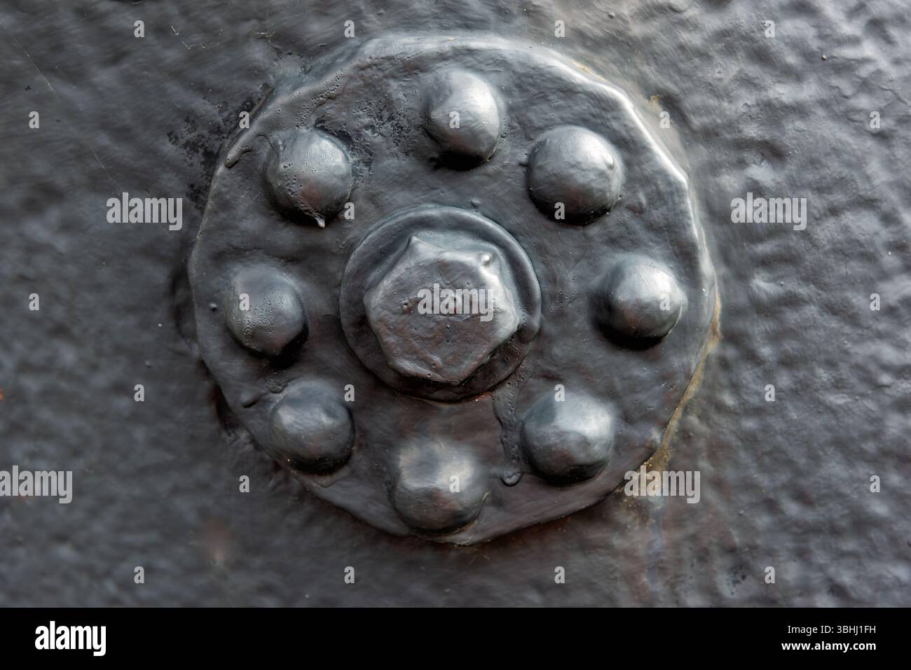 Der strukturierte Metallbolzen mit Nieten hebt sich von einer dunklen Industrieoberfläche ab, die von natürlichem Licht beleuchtet wird und sein Design und seine Struktur unterstreicht. Stockfoto